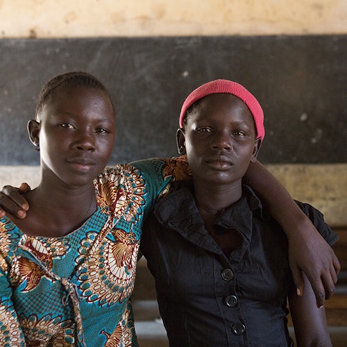 deux jeunes filles ougandaises se tiennent par le bras et sourient à la caméra dans une salle de classe.