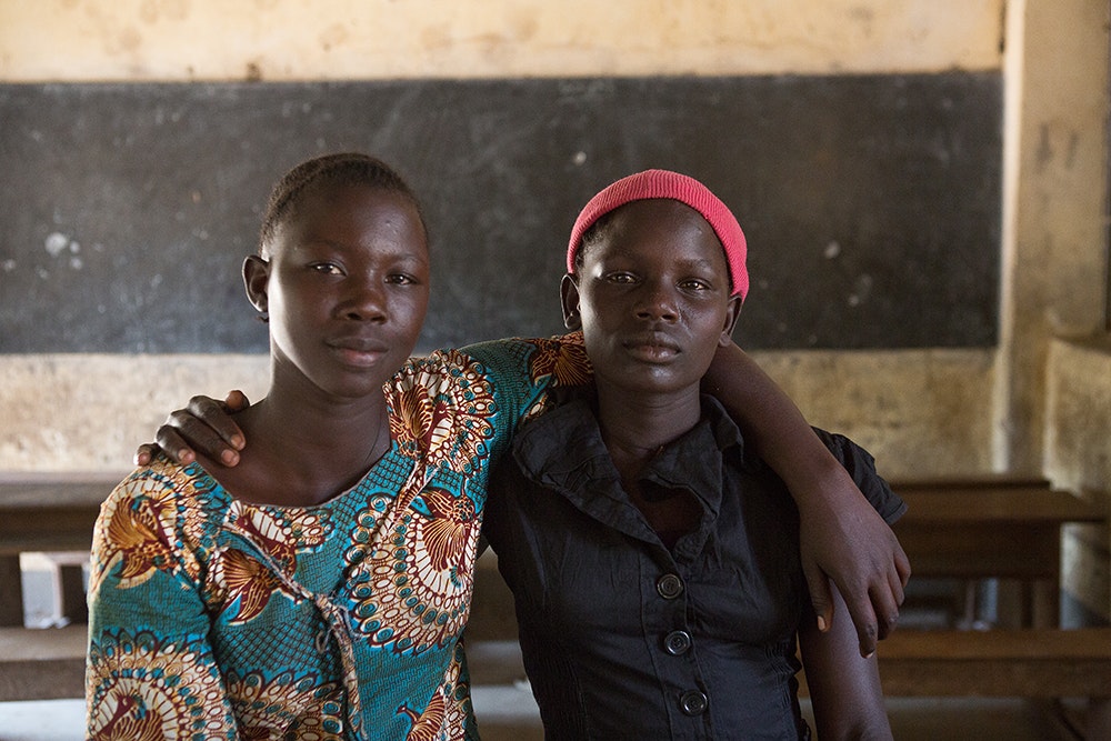 two Uganda girls with hand on each other's arm and smile to the camera inside the classroom