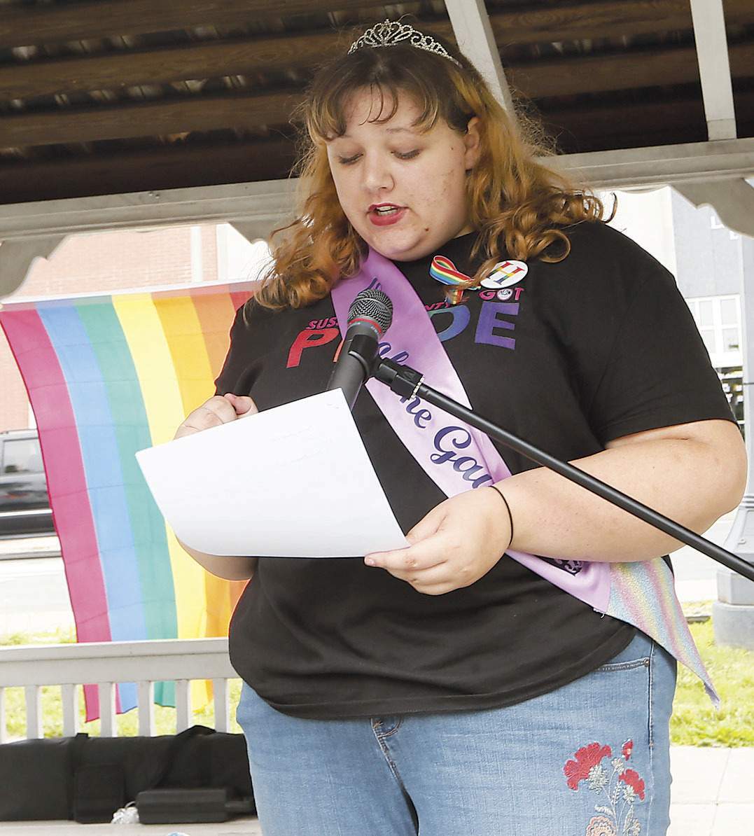 (half body hot) Zoe Heath with her pride shirt and right hand holding a paper and speaking in front of the standing microphone