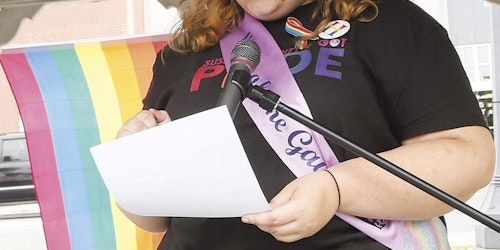 Zoe Heath usando uma camiseta com a palavra “Pride” (orgulho), segurando um papel com a mão direita e falando ao microfone (foto de perto)