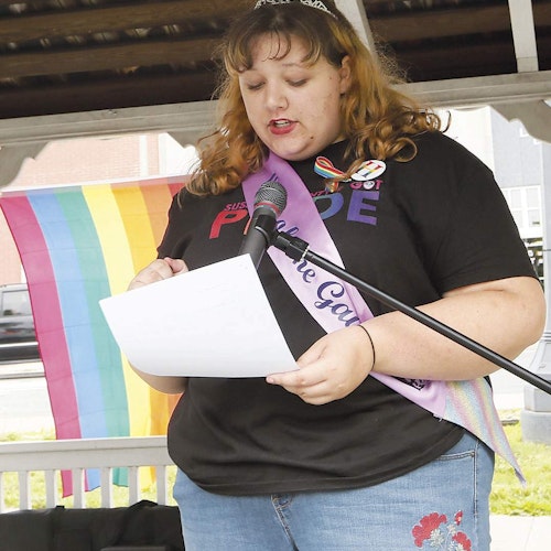 (meio corpo quente) Zoe Heath com a sua camisa de orgulho e mão direita segurando um papel e falando em frente ao microfone de pé