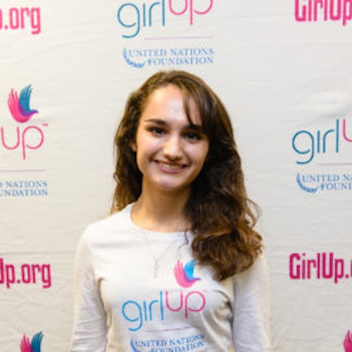 Alex Leone, consultora adolescente de 2013-2014 (foto de perto, um pouco desfocada). Uma adolescente sorridente olhando para a câmera, tendo uma parede com “girlup.org” no plano de fundo. Ela está usando a camiseta toda branca da Girl Up