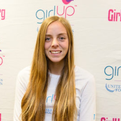 Alexis Kallen, consultora adolescente de 2013-2014 (foto de perto, um pouco desfocada). Uma adolescente sorridente olhando para a câmera, tendo uma parede com “girlup.org” no plano de fundo. Ela está usando a camiseta toda branca da Girl Up