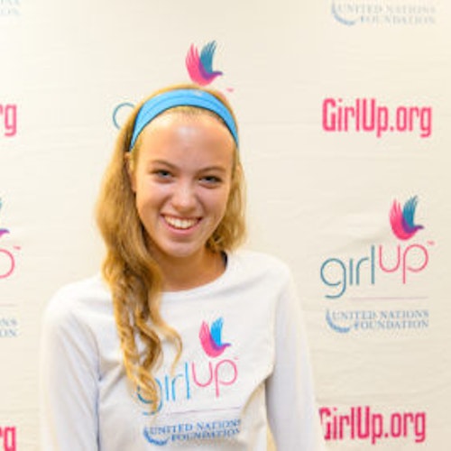 Amanda Hart_Consejera adolescente 2013-2014 (retrato en primer plano, fotografía un poco borrosa); una adolescente con la camiseta blanca de Girl Up, sonriendo a la cámara, con el cartel de girlup.org de fondo.