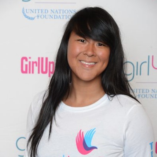 Amy Gong Liu, consultora adolescente de 2014-2015 (foto de perto). Uma adolescente sorridente olhando para a câmera, tendo uma parede com “girlup.org” no plano de fundo. Ela está usando a camiseta toda branca da Girl Up