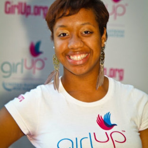 Angelique Gaston_家乡： 美国华盛顿特区_2011-2012 届（即第二届）青年顾问（近距离头像照），照片中的她穿着白色 Girl Up T 恤，面对镜头微笑，照片背景为 girlup.org 活动展板