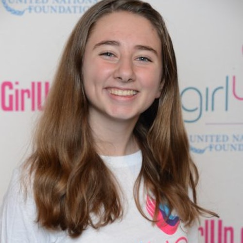 Anna McGuire, consultora adolescente de 2014-2015 (foto de perto). Uma adolescente sorridente olhando para a câmera, tendo uma parede com “girlup.org” no plano de fundo. Ela está usando a camiseta toda branca da Girl Up
