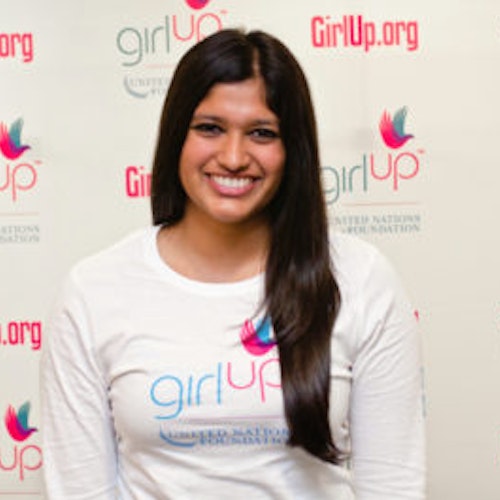 Archana Somasegar, consultora adolescente de 2012-2013 (foto de perto, um pouco desfocada). Uma adolescente sorridente olhando para a câmera, tendo uma parede com “girlup.org” no plano de fundo. Ela está usando a camiseta toda branca da Girl Up