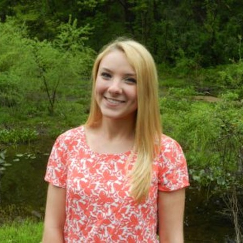 Becca Bean, consultora adolescente de 2016-2017 (foto de meio-corpo, desfocada), sorridente olhando para a câmera, tendo o verde das plantas como plano de fundo
