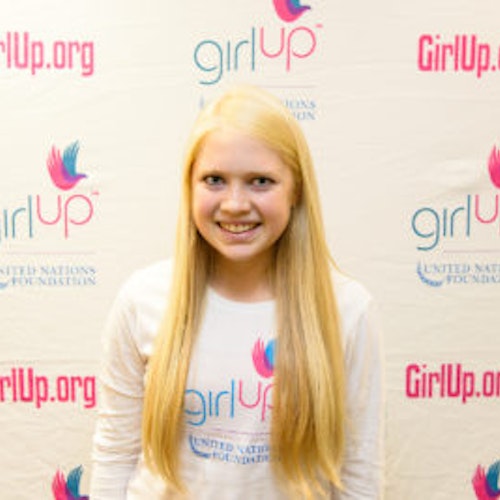 Carly Bandt_2013-2014 Teen Advisor (photo rapprochée, un peu floue) une adolescente portant sa chemise blanche girl up avec son visage souriant face à l'appareil photo, et en arrière-plan le conseil d'administration de girlup.org.