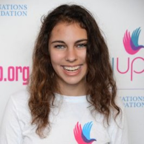 Celia Buckman, codirectora_Consejeras adolescentes 2015-2016 (retrato en primer plano, fotografía un poco borrosa); una adolescente con la camiseta blanca de Girl Up, sonriendo a la cámara, con el cartel de girlup.org de fondo.