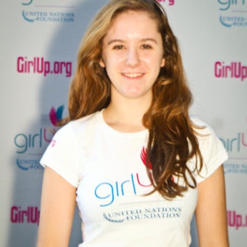 Charlotte May_家乡： 美国纽约州布朗克斯维尔_2011-2012 届（即第二届）青年顾问（近距离头像照），照片中的她穿着白色 Girl Up T 恤，面对镜头微笑，照片背景为 girlup.org 活动展板