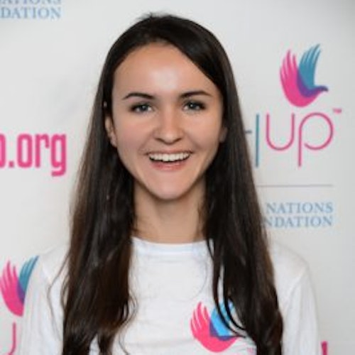 Claire Brito, copresidente, consultora adolescente de 2015-2016 (foto de perto), sorridente olhando para a câmera, tendo uma parede com “girlup.org” no plano de fundo. Ela está usando a camiseta toda branca da Girl Up