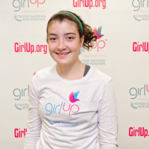 Delia Friel_Grupo de Consejeras adolescentes 2012-2013 (retrato en primer plano, fotografía un poco borrosa); una adolescente con la camiseta blanca de Girl Up, sonriendo a la cámara, con el cartel de girlup.org de fondo.