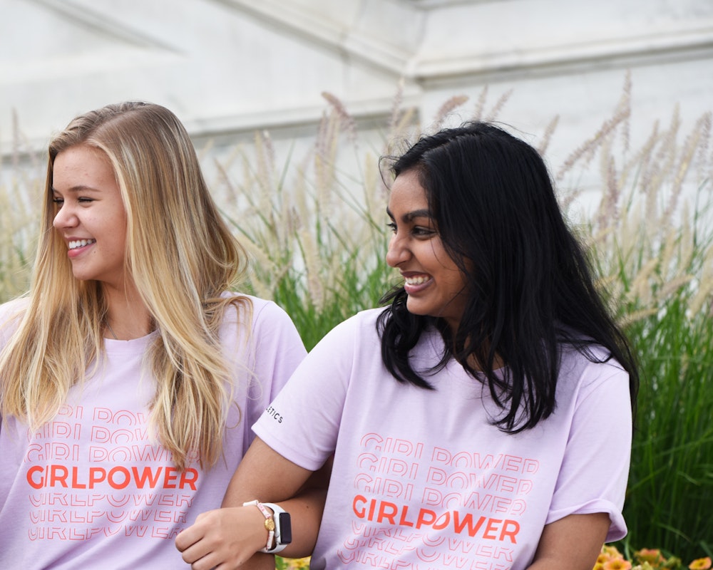 3 chicas mirando cada una y con la mano alrededor del brazo de la otra, llevando una camiseta &quot;girl power&quot;.