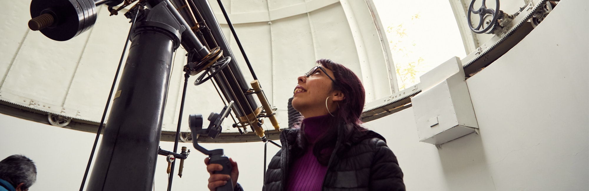 Mulher olhando para cima, à frente de um telescópio enorme