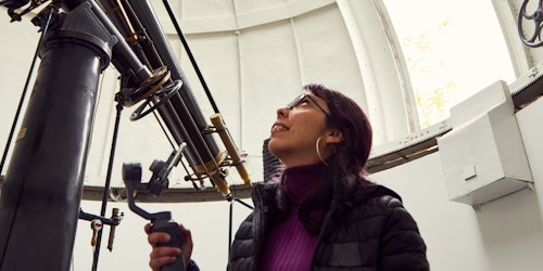 Mulher olhando para cima, à frente de um telescópio enorme