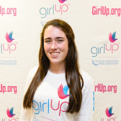 Emma Knoll_Consejera adolescente 2013-2014 (retrato en primer plano, fotografía un poco borrosa); una adolescente con la camiseta blanca de Girl Up, sonriendo a la cámara, con el cartel de girlup.org de fondo.