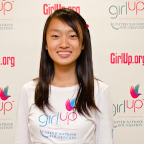 Eva (YingYing) Shang, consultora adolescente de 2012-2013 (foto de perto, um pouco desfocada). Uma adolescente sorridente olhando para a câmera, tendo uma parede com “girlup.org” no plano de fundo. Ela está usando a camiseta toda branca da Girl Up