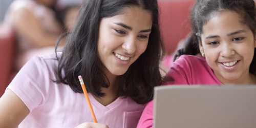 two girls are smiling and writing down notes and sharing a laptop