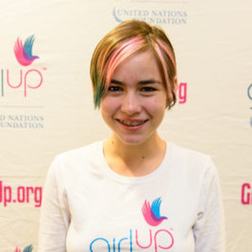 Giuliana Gabellini, consultora adolescente de 2013-2014 (foto de perto, um pouco desfocada). Uma adolescente sorridente olhando para a câmera, tendo uma parede com “girlup.org” no plano de fundo. Ela está usando a camiseta toda branca da Girl Up