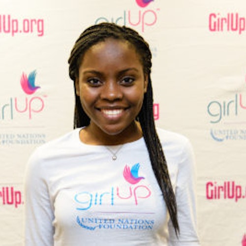 Gloria Samen, consultora adolescente de 2013-2014 (foto de perto, um pouco desfocada). Uma adolescente sorridente olhando para a câmera, tendo uma parede com “girlup.org” no plano de fundo. Ela está usando a camiseta toda branca da Girl Up