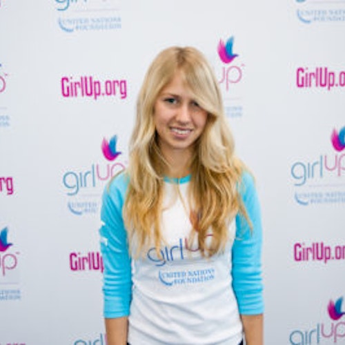 Hadley Walsh_A turma fundadora da Teen Advisors(ângulo estreito, mas não imagem clara) uma adolescente com a sua camisa comprida azul com a cara sorridente virada para a câmara, e o fundo é o quadro girlup.org