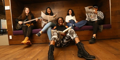 (Fotografía a distancia) En la fotografía, cinco chicas de diferentes etnias sostienen libros (sentadas adelante en el piso) y otras, una guitarra y un periódico (sentadas en el sofá de atrás).