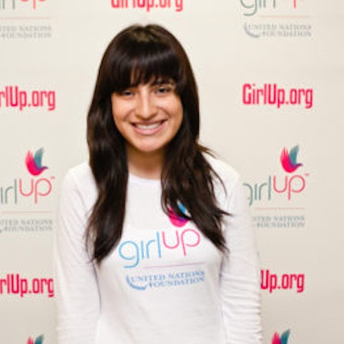 Ines Renique_家乡： 美国纽约州纽约市_2012-2013 届青年顾问（近距离头像照，画面有点模糊），照片中的她穿着白色 Girl Up T 恤，面对镜头微笑，照片背景为 girlup.org 活动展板