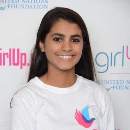 Ishana Nigam, consultora adolescente de 2014-2015 (foto de perto). Uma adolescente sorridente olhando para a câmera, tendo uma parede com “girlup.org” no plano de fundo. Ela está usando a camiseta toda branca da Girl Up