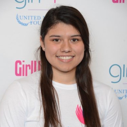 Janet Diaz, consultora adolescente de 2014-2015 (foto de perto). Uma adolescente sorridente olhando para a câmera, tendo uma parede com “girlup.org” no plano de fundo. Ela está usando a camiseta toda branca da Girl Up