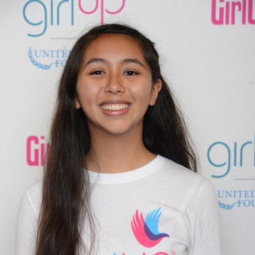 Jessica Bishai_Consejera adolescente 2014-2015 (retrato en primer plano); una adolescente con la camiseta blanca de Girl Up, sonriendo a la cámara, con el cartel de girlup.org de fondo.