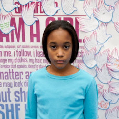 Jillian Medina,a classe fundadora da Teen Advisors(ângulo estreito, mas não claro) uma menina adolescente vestindo a sua camisa comprida azul de frente para a câmara, e o fundo é o quadro girlup.org