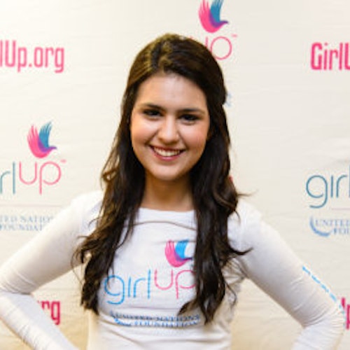 Julia Sumouske, consultora adolescente de 2013-2014 (foto de perto, um pouco desfocada). Uma adolescente sorridente olhando para a câmera, tendo uma parede com “girlup.org” no plano de fundo. Ela está usando a camiseta toda branca da Girl Up