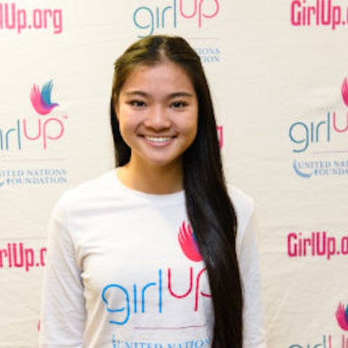 Kaitlin Hung_Consejera adolescente 2013-2014 (retrato en primer plano, fotografía un poco borrosa); una adolescente con la camiseta blanca de Girl Up, sonriendo a la cámara, con el cartel de girlup.org de fondo.