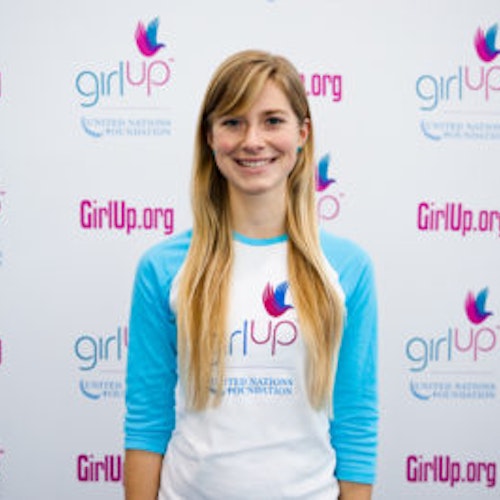 Karina Jougla_ A classe fundadora dos Teen Advisors(ângulo estreito, mas não claro) uma adolescente com a sua camisa comprida azul com o seu rosto sorridente virado para a câmara, e o fundo é o quadro girlup.org