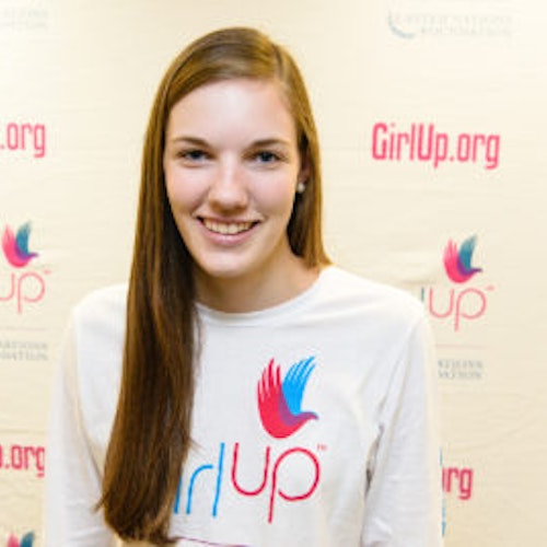 Kate McCollum, consultora adolescente de 2013-2014 (foto de perto, um pouco desfocada). Uma adolescente sorridente olhando para a câmera, tendo uma parede com “girlup.org” no plano de fundo. Ela está usando a camiseta toda branca da Girl Up
