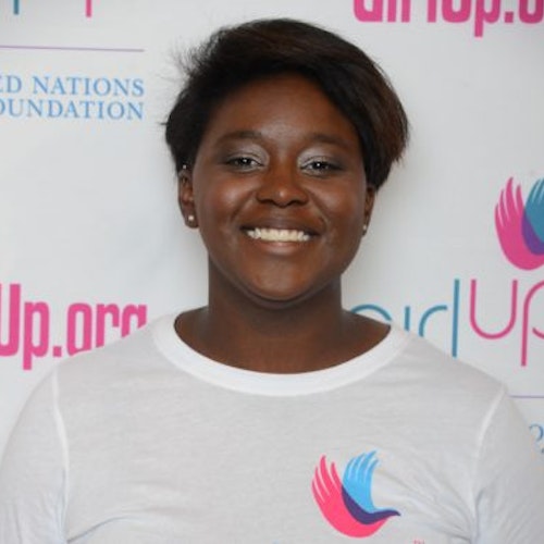 Kennede Reese, consultora adolescente de 2014-2015 (foto de perto). Uma adolescente sorridente olhando para a câmera, tendo uma parede com “girlup.org” no plano de fundo. Ela está usando a camiseta toda branca da Girl Up
