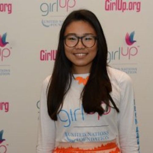 Kyung Mi Lee, copresidente, consultora adolescente de 2016-2017 (foto de meio-corpo, desfocada), sorridente olhando para a câmera, tendo uma parede com “girlup.org” no plano de fundo. Ela está usando a camiseta toda branca da Girl Up