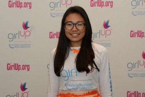 Kyung Mi Lee, Co-Chair_2016-2017 Teen Advisors (wider angle half-body , blurry pciture) wearing her girl up white shirt with her smiley face facing the camera, and background is girlup.org board