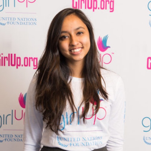 Lavanya Singh, codirectora, Consejeras adolescentes de 2017-2018 (retrato), sonriendo a la cámara con su camiseta blanca de Girl Up y con el cartel de girlup.org de fondo.