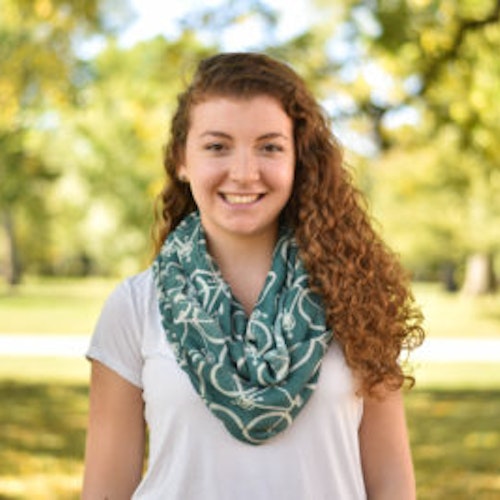 Leah Adelman, consultora adolescente de 2016-2017 (foto de meio-corpo em ângulo amplo, desfocada) sorridente olhando para a câmera, tendo o verde das plantas como plano de fundo