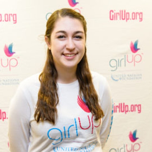 Lindsay Schrier, consultora adolescente de 2013-2014 (foto de perto, um pouco desfocada). Uma adolescente sorridente olhando para a câmera, tendo uma parede com “girlup.org” no plano de fundo. Ela está usando a camiseta toda branca da Girl Up