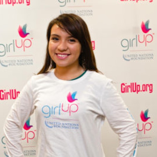 Martha Zuniga_家乡： 美国伊利诺伊州芝加哥_2012-2013 届青年顾问（近距离头像照，画面有点模糊），照片中的她穿着白色 Girl Up T 恤，面对镜头微笑，照片背景为 girlup.org 活动展板