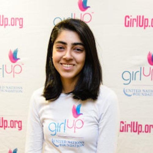 Mehar Gujral_Consejera adolescente 2013-2014 (retrato en primer plano, fotografía un poco borrosa); una adolescente con la camiseta blanca de Girl Up, sonriendo a la cámara, con el cartel de girlup.org de fondo.