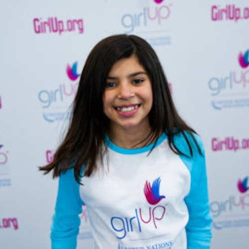 Mia Gutierrez, A turma fundadora da Teen Advisors (ângulo estreito, mas sem imagem nítida) uma adolescente com a sua camisa comprida azul com o seu rosto sorridente virado para a câmara, e o fundo é o quadro girlup.org