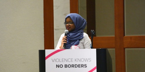 Consultora adolescente Munira Alimire segurando um microfone e falando atrás de um púlpito. Na frente do púlpito há uma placa com a frase “Violence knows no borders” (A violência não tem fronteiras)