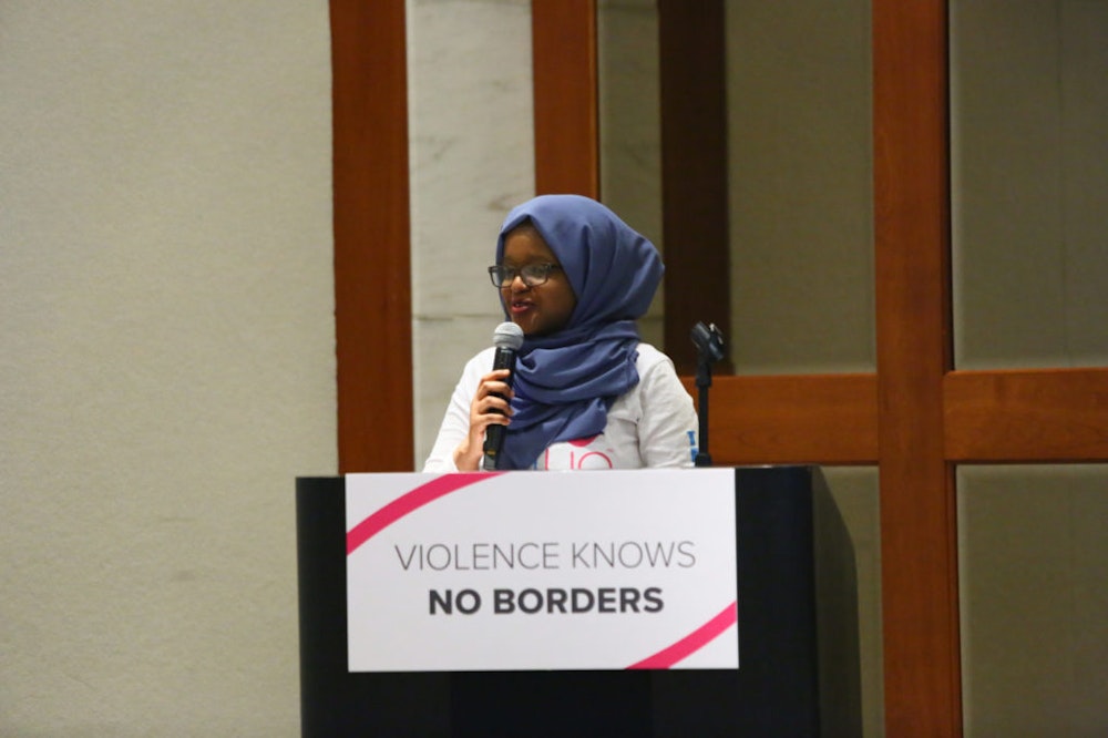 Munira Alimire teen adviser holding a mic and speaking behind the podium, in the front of the podium a sign "violence knows no borders"
