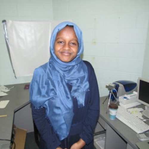 Munira Alimire, consultora adolescente de 2017-2018 (foto de meio-corpo, desfocada) tendo um laboratório como plano de fundo. Ela está usando um hijab azul