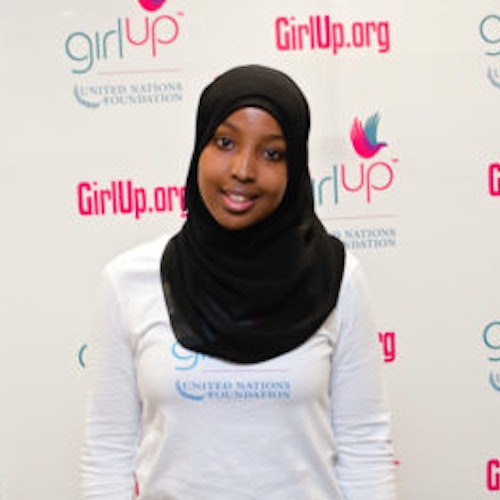 Munira Khalif_家乡： 美国明尼苏达州弗里德利_2012-2013 届青年顾问（近距离头像照，画面有点模糊），照片中的她穿着白色 Girl Up T 恤，面对镜头微笑，照片背景为 girlup.org 活动展板
